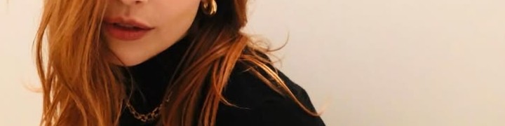 Fotografia de uma mulher ruiva com roupa preta e colares, enquadrada parcialmente no rosto e no tronco.