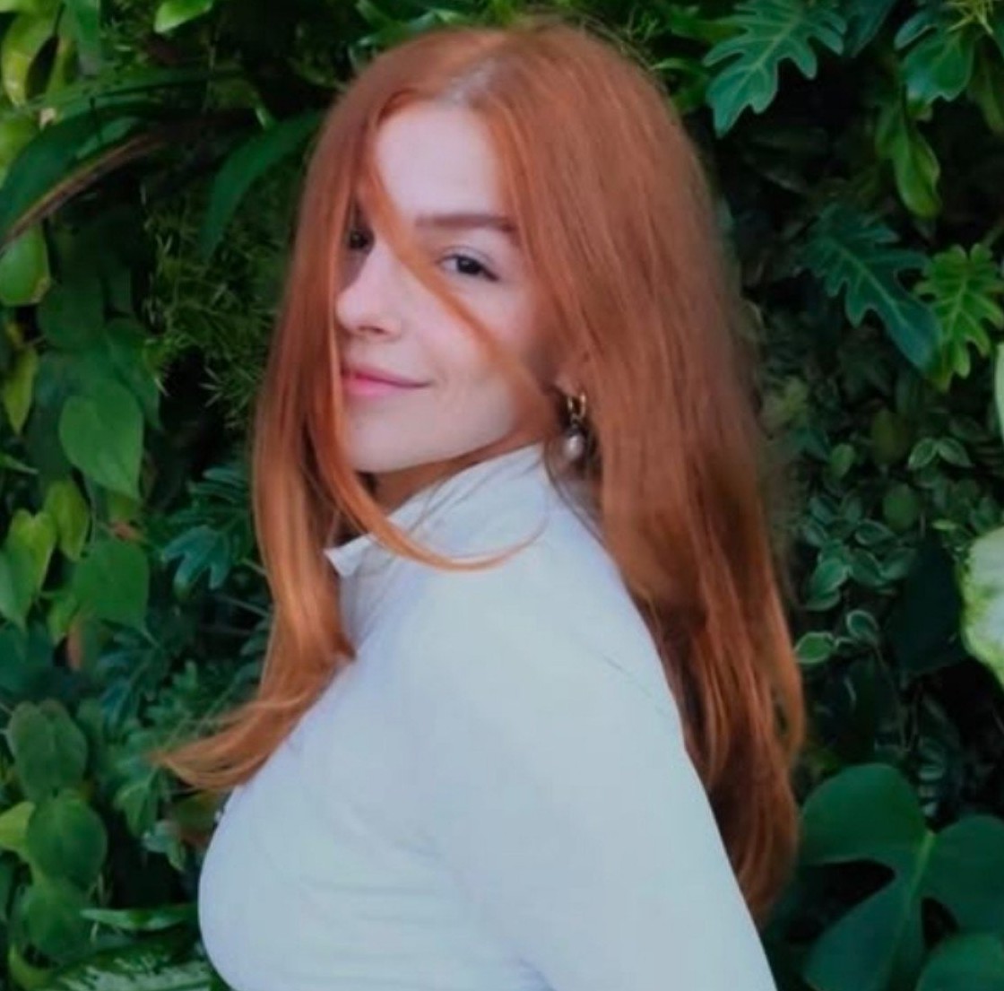 Retrato de uma mulher ruiva com roupa clara, olhando por cima do ombro diante de folhagens.
