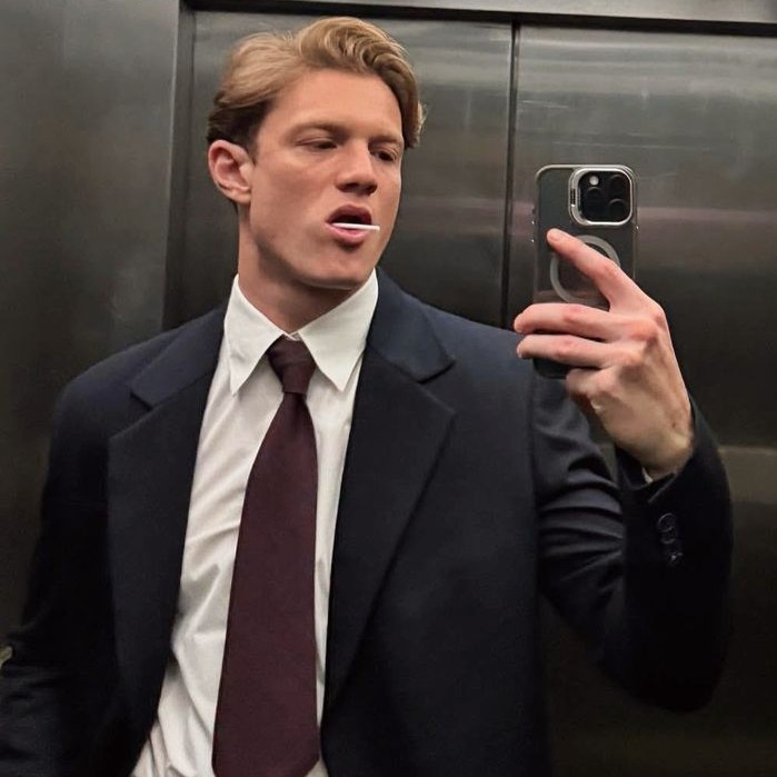 Retrato de um homem de terno e gravata segurando um celular diante de um espelho.