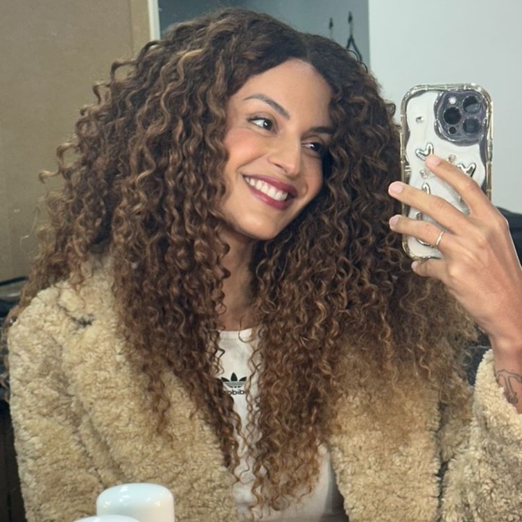 Retrato de uma mulher de cabelos cacheados sorrindo e segurando um celular.
