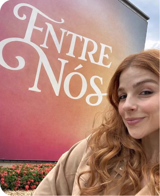 Eduarda Fragoso diante de um painel com o título Entre Nós, olhando para a câmera.
