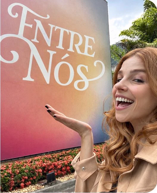 Eduarda Fragoso sorrindo e apresentando com a mão um painel com o título Entre Nós.