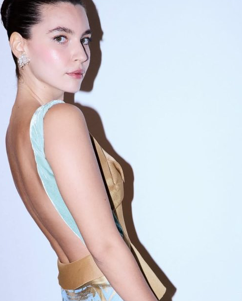 Mulher de cabelos escuros presos olhando sobre o ombro, com vestido dourado e detalhe azul-claro nas costas.